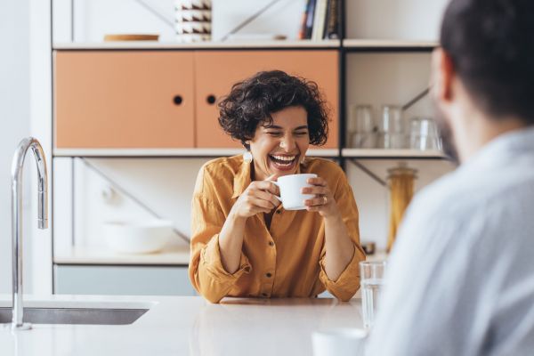 Woman leaning on kitchen counter while holding a cup of coffee and laughing